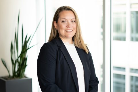 A woman in a black blazer and white top stands indoors near a large window, with a potted plant visible in the background.
