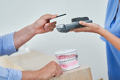 A person pays with a credit card using a card reader at a dental office reception, with a model of teeth on the counter.