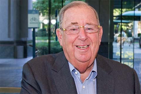 An older man wearing glasses and a suit jacket smiles at the camera while standing indoors near large glass windows.