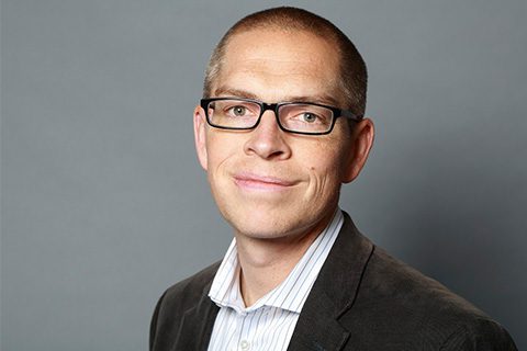 A man with short hair and glasses wears a striped shirt and dark blazer, standing in front of a plain gray background.