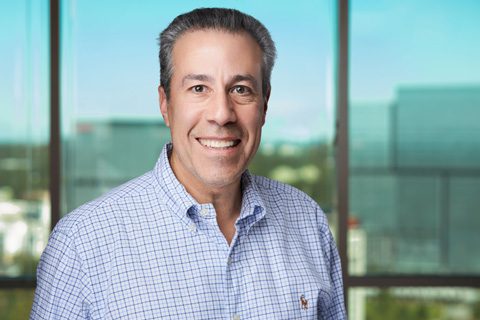 A man in a light blue checkered shirt smiles at the camera, standing indoors with large windows and an office building visible in the background.