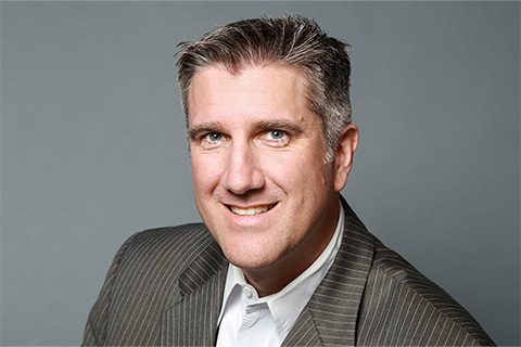 A man with short gray hair wearing a pinstripe suit jacket and white shirt smiles at the camera against a plain gray background.
