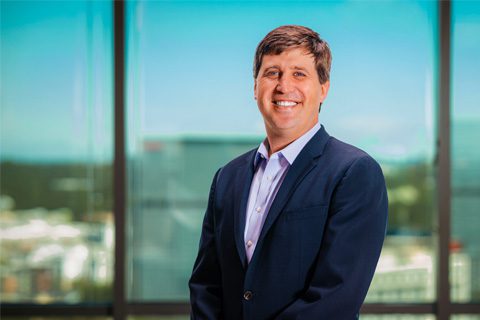 A man in a dark suit smiles, standing indoors in front of large windows with a cityscape blurred in the background.