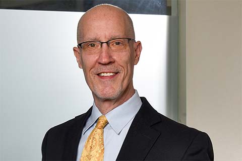 A middle-aged man with glasses, a suit, and a yellow patterned tie is smiling at the camera in an indoor setting.