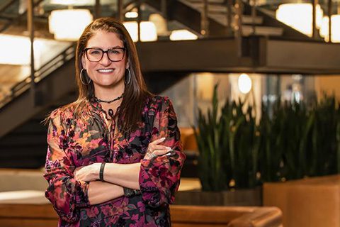 A woman with long brown hair and glasses, wearing a floral blouse, stands with arms crossed in a modern indoor space with a staircase and plants in the background.
