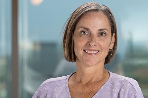 A woman with short brown hair and a light purple patterned top stands indoors, smiling at the camera with a blurred cityscape in the background.
