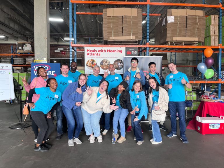 Group of volunteers in matching blue Aprio tshirts pose together in a warehouse