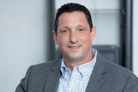 A man with short dark hair, wearing a gray blazer and a checked shirt, smiles at the camera in an office setting.