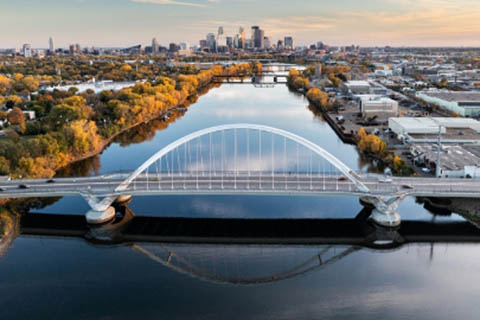 A modern white arch bridge spans a calm river with a city skyline and autumn trees in the background.