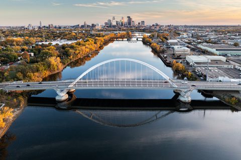 A white arched bridge spans a calm river with city buildings and trees lining the banks, under a partly cloudy sky at sunset.