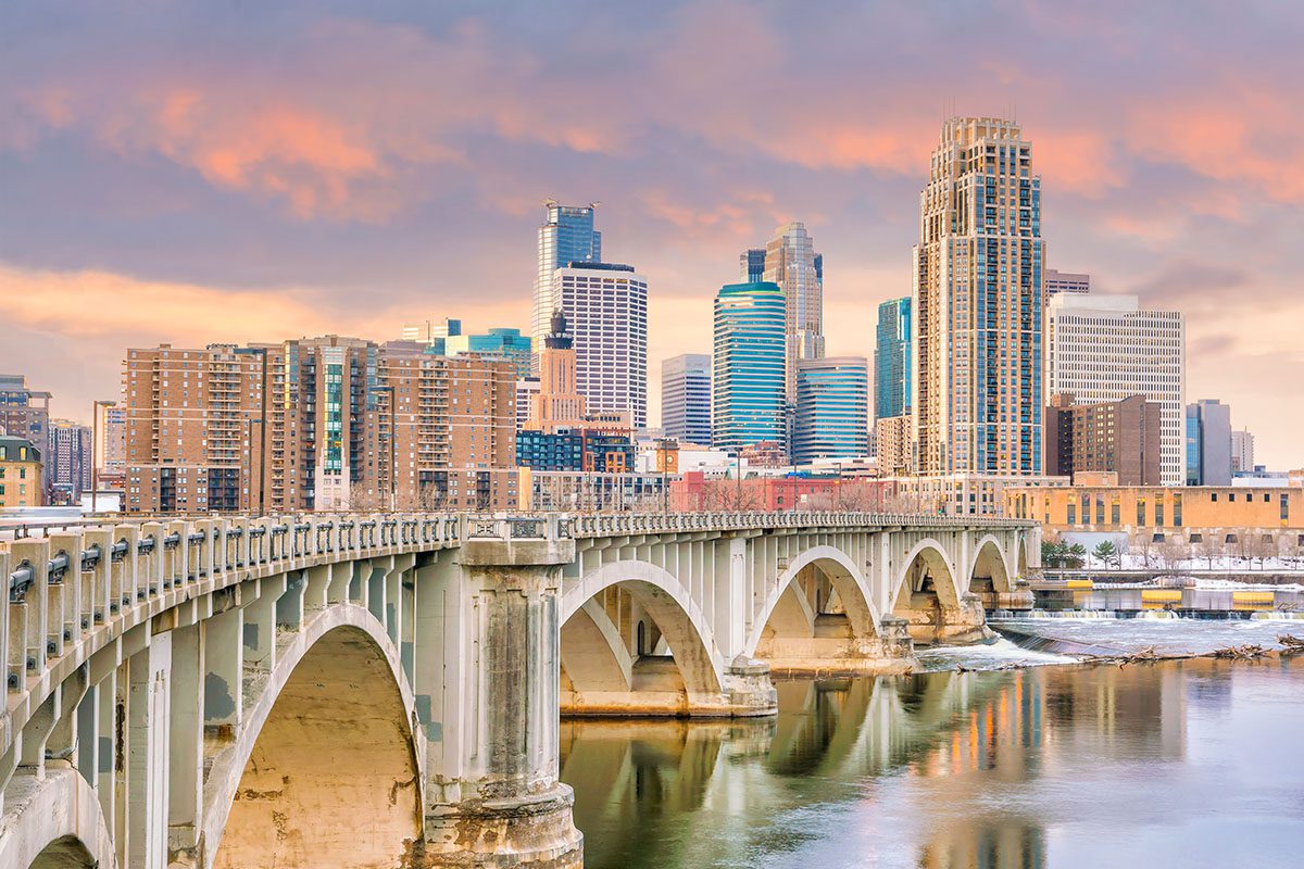 Stone arch bridge over a river with downtown Minneapolis skyscrapers in the background under a partly cloudy sunset sky.