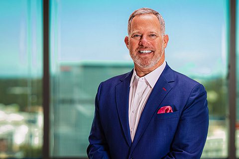 A man in a blue suit with a pink pocket square stands indoors in front of large windows overlooking a cityscape, smiling at the camera.
