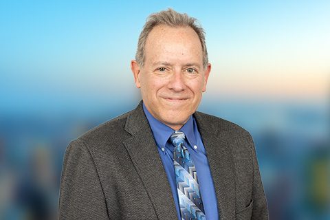 A middle-aged man in a suit, blue shirt, and patterned tie stands in front of a blurred, cityscape background.