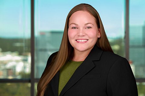 A woman with long straight brown hair, wearing a black blazer and green top, smiles while standing in front of a window overlooking a cityscape.