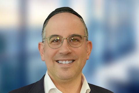 A man with short hair, wearing glasses, a dark suit jacket, and a black kippah, smiling in front of a blurred blue background.