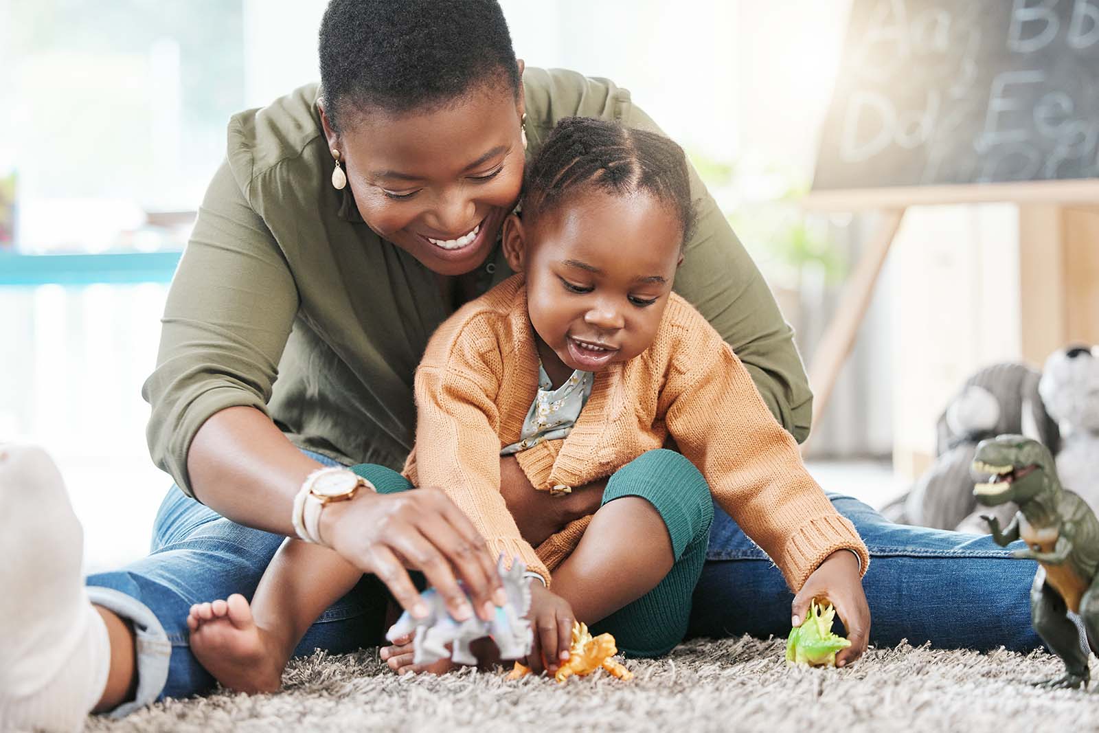 An adult and a young child sit on the floor, smiling and playing with toy animals together in a brightly lit room.