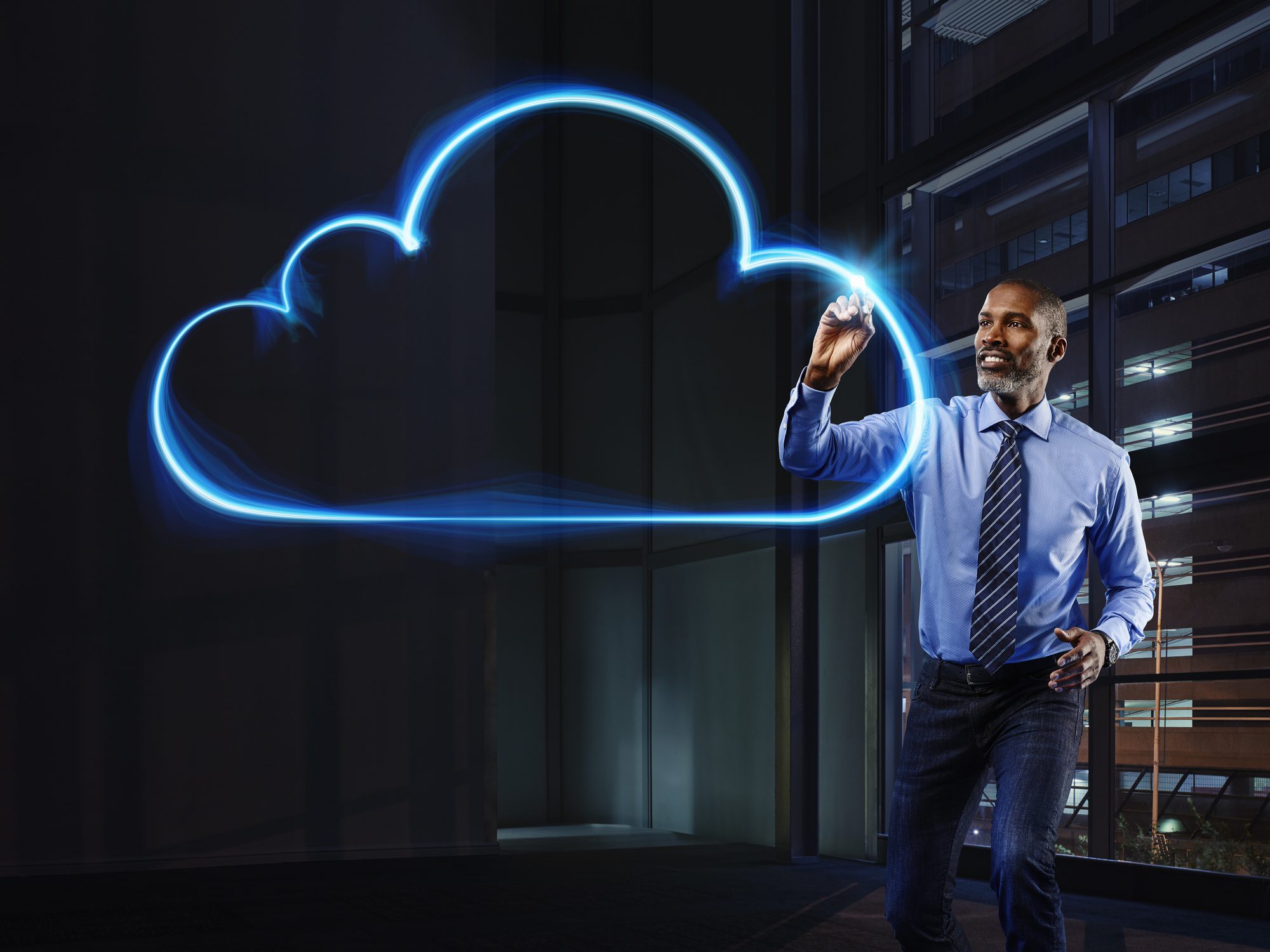 A man in business attire stands indoors, drawing a glowing blue cloud outline in the air, symbolizing cloud computing or digital technology.