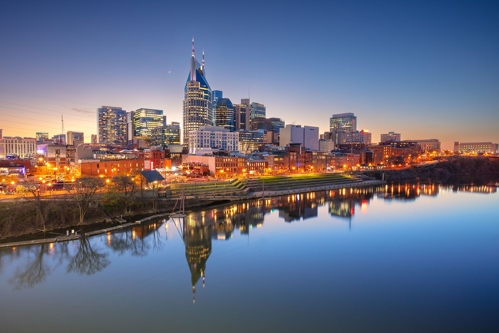Nashville city skyline at dusk with buildings lit up and their reflections visible in the calm river in the foreground.