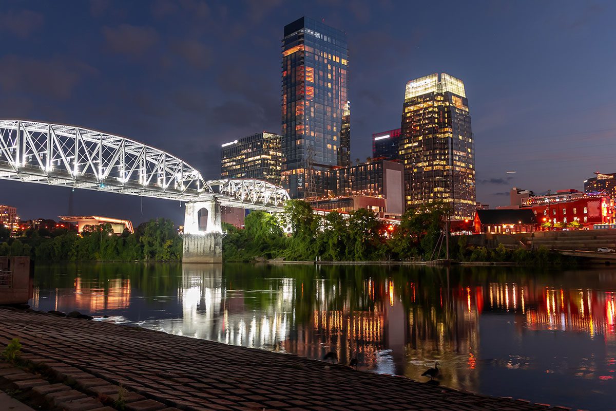 City skyline at dusk with illuminated buildings and a lit bridge reflecting on a calm river, viewed from a cobblestone riverbank.