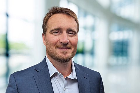 A man with short brown hair and a beard, wearing a blue suit jacket and white shirt, stands indoors in front of large windows.