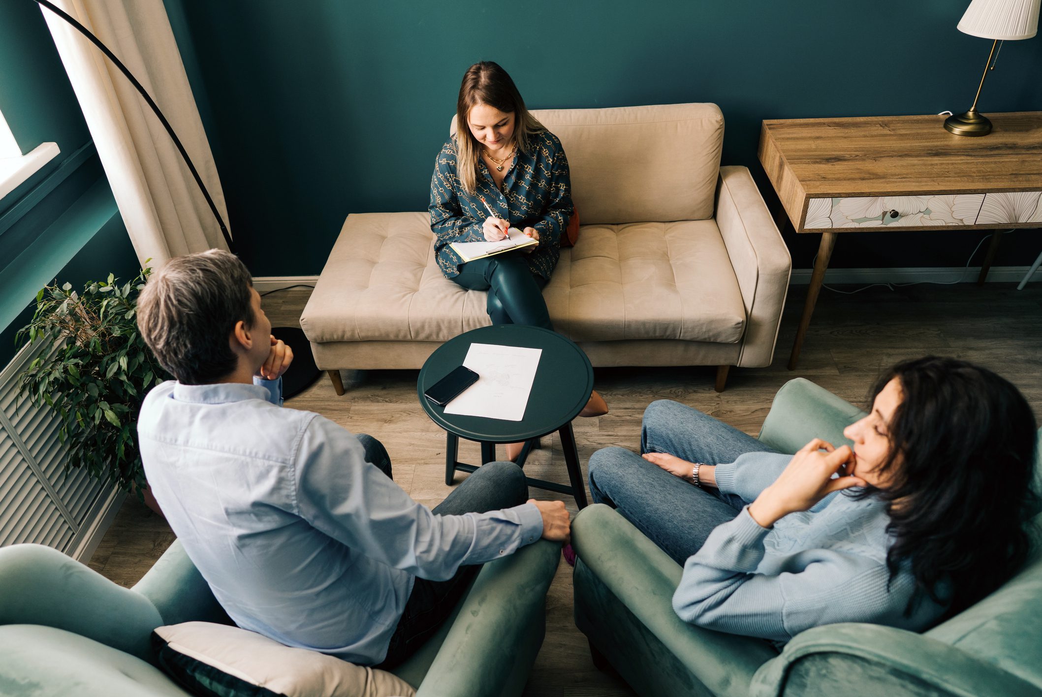Three people sit in a living room, with two facing a professional who is taking notes on a clipboard, suggesting a counseling or therapy session.