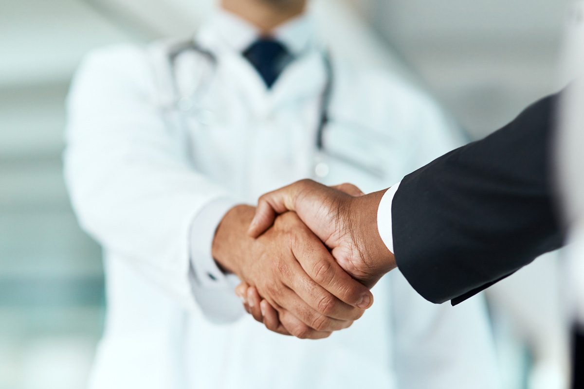 A doctor in a white coat and a person in a suit shake hands, suggesting a professional agreement or partnership.