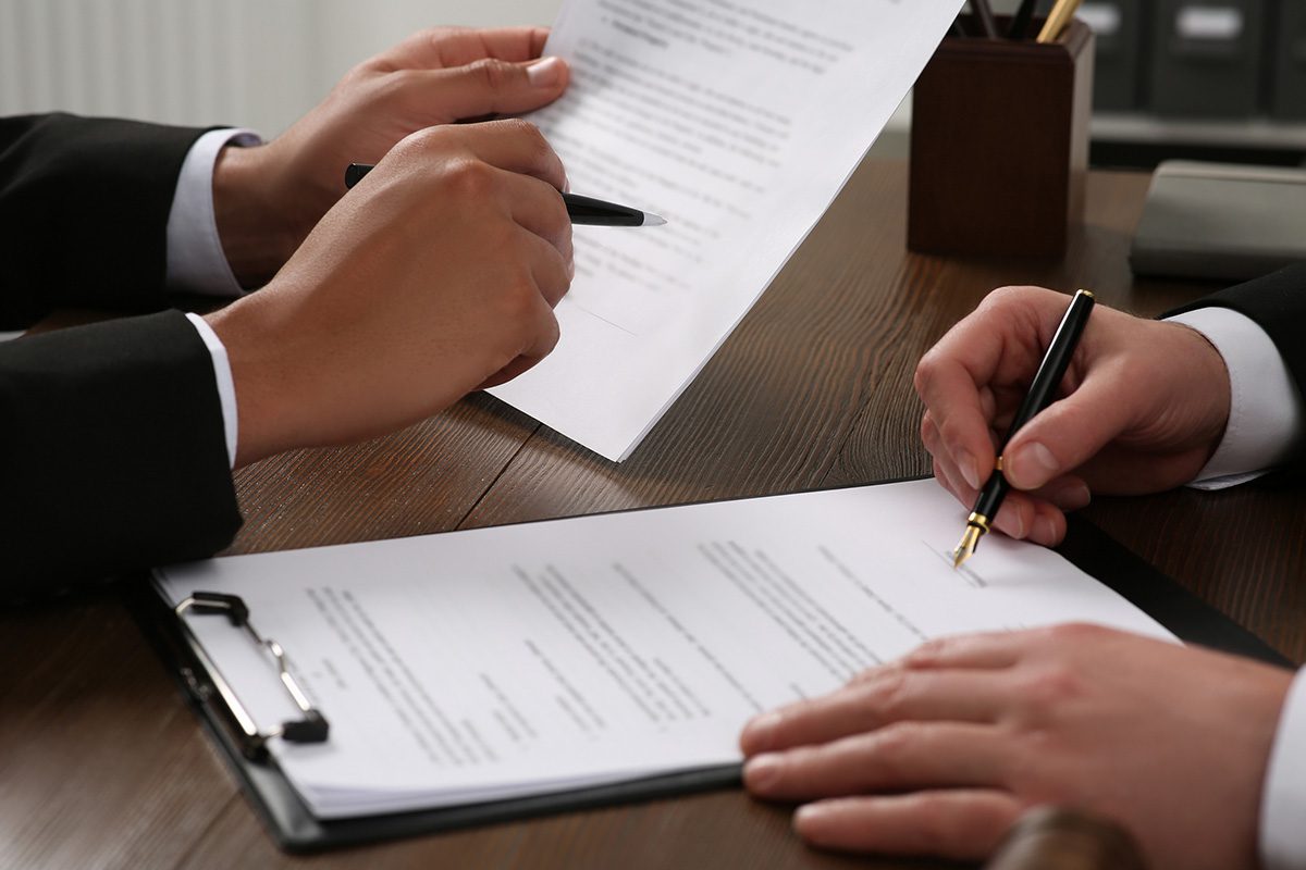Two people in business attire review and sign documents on a wooden desk, with pens in hand and paperwork spread out in front of them.