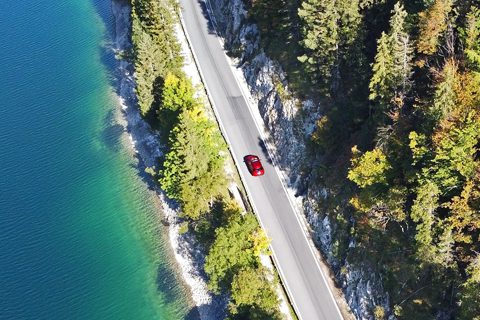 A red car drives on a road running beside a turquoise lake and dense forest, viewed from above.