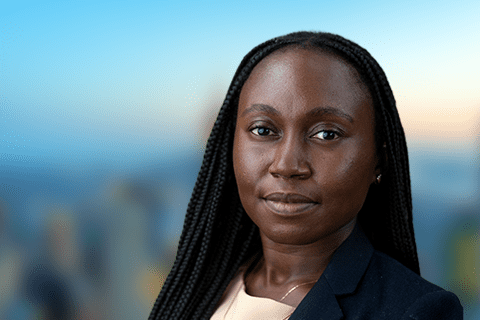 A woman with long braided hair wearing a dark blazer and light top faces the camera with a neutral expression; the background is blurred cityscape.