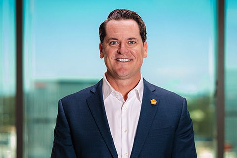 Dark haired man in a blazer smiles in front of a bright window with a blurred city view