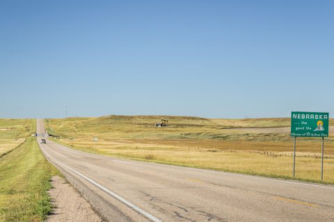 A paved highway stretches through grassy plains under a clear sky, with a green road sign reading Nebraska, Home of Arbor Day on the right side of the road.