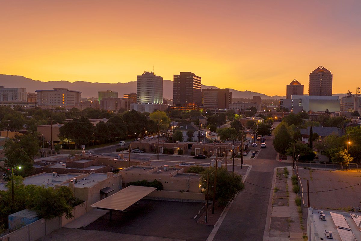 City skyline at sunset with tall buildings, residential houses, and mountains in the background under an orange sky.