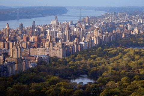 Aerial view of New York City skyline with tall buildings alongside Central Park, with the Hudson River and a distant bridge in the background.