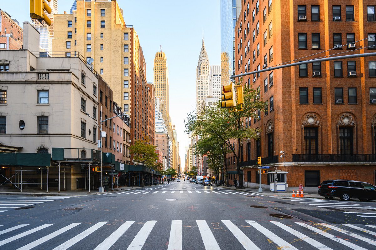 A wide, empty city street lined with tall buildings, crosswalks, and traffic lights, with the Chrysler Building visible in the background under a clear sky.