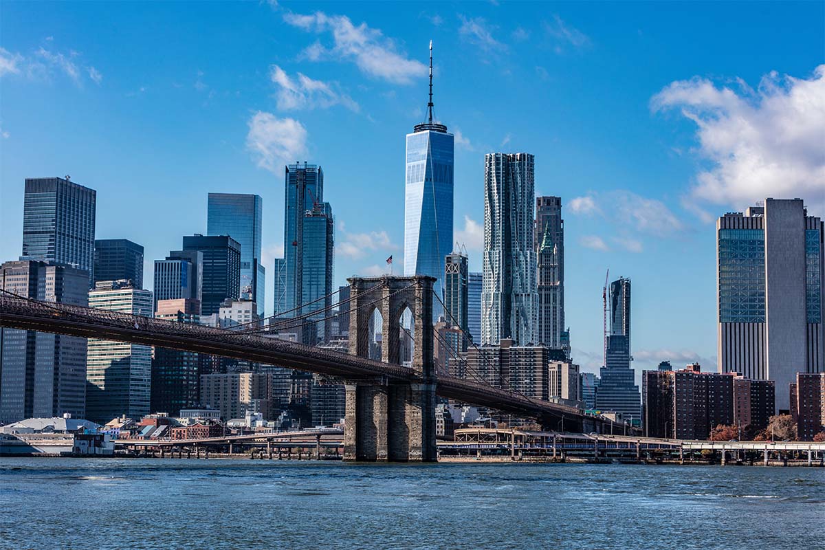 View of the Brooklyn Bridge spanning the East River with the Manhattan skyline, including the One World Trade Center, under a blue sky.