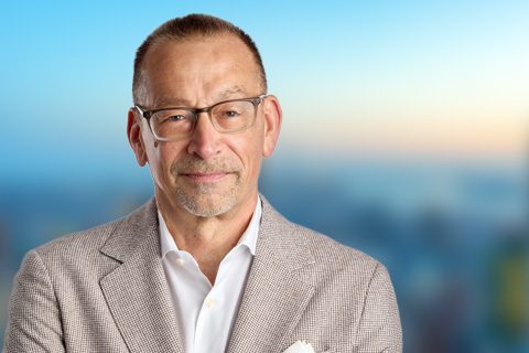 A middle-aged man wearing glasses and a light-colored blazer stands in front of a blurred outdoor background.