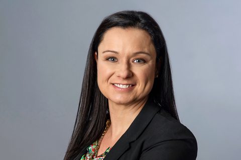 Woman with long straight dark hair wearing a black blazer and patterned top, smiling at the camera against a plain gray background.