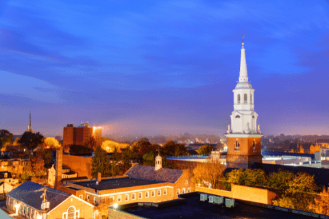 View of a town at dusk featuring a prominent white church steeple and brick buildings under a blue sky with scattered clouds.