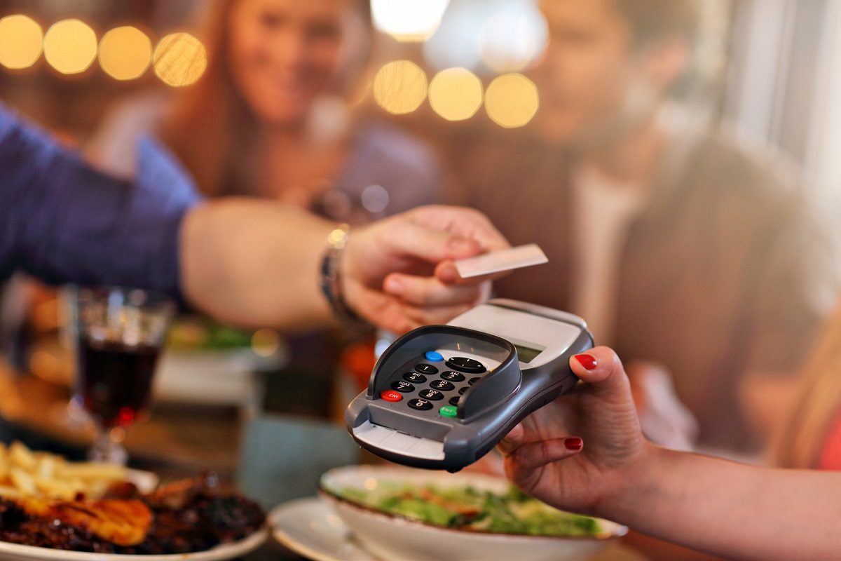 A person hands a credit card to another person holding a card reader at a restaurant table with food and drinks.