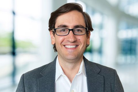 A man with glasses and brown hair, wearing a white shirt and gray blazer, smiles at the camera in a bright, modern office setting.