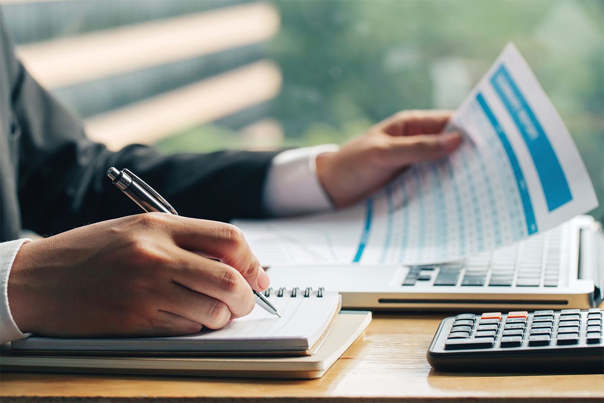 Person in business attire writes in a notebook while holding a report, with a laptop and calculator on the desk.