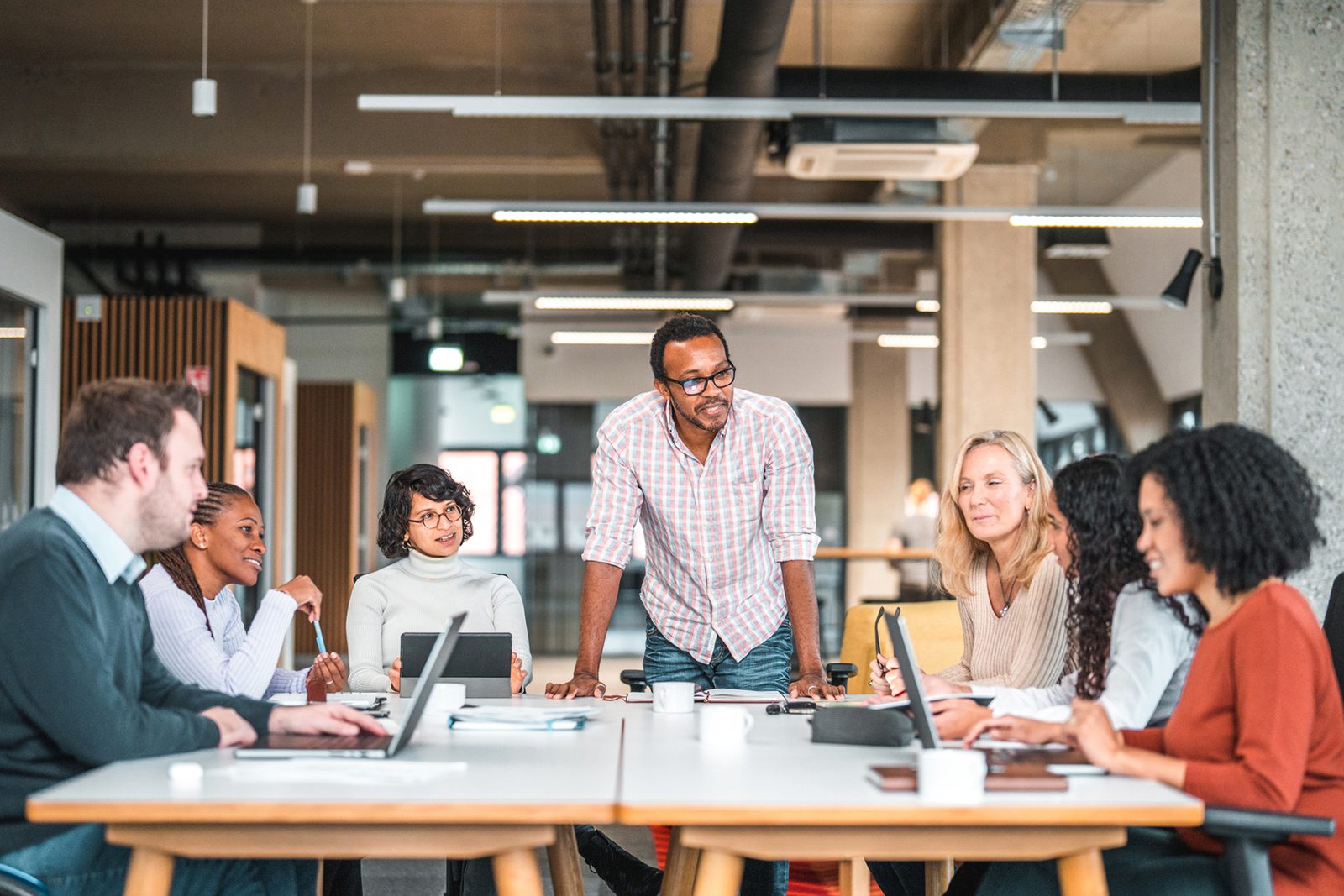 A group of six people sit around a conference table with laptops, while one person stands and speaks in a modern office setting.