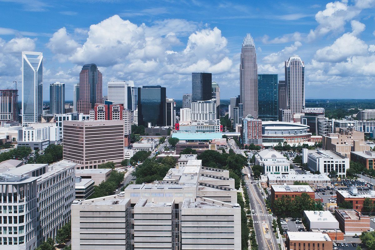 Aerial view of a city skyline with tall modern skyscrapers, mid-rise buildings, and tree-lined streets under a partly cloudy sky.