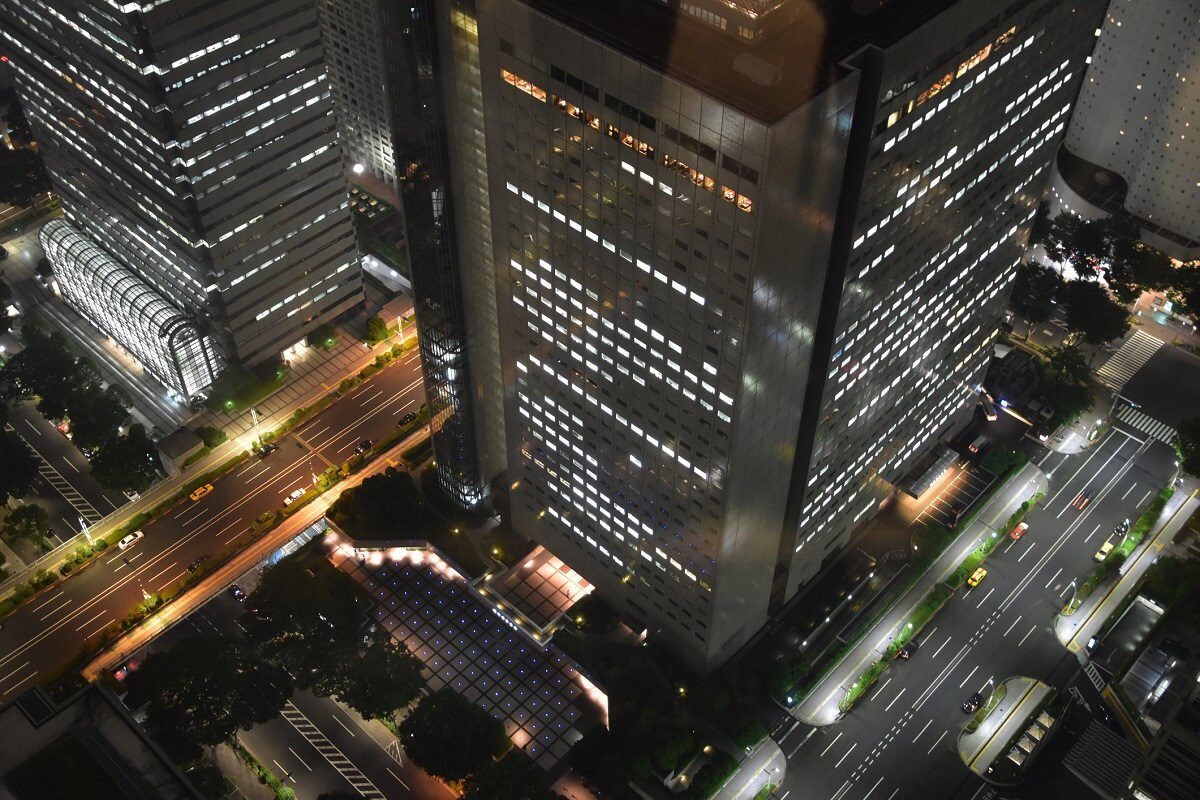Aerial night view of tall office buildings with illuminated windows, busy city streets, and moving traffic in an urban downtown area.
