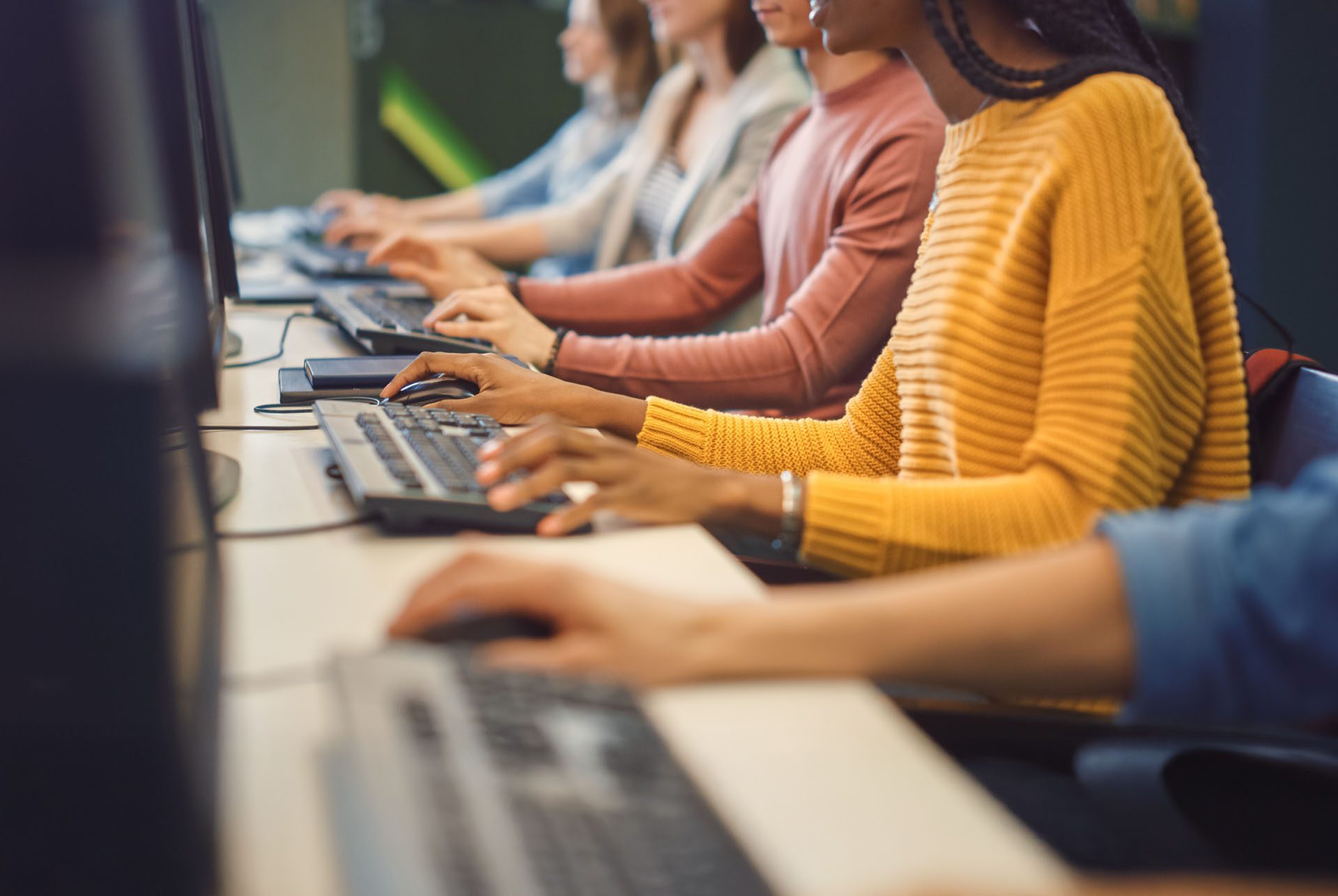 Several people sit in a row at desks, typing on desktop computers in a classroom or office setting.