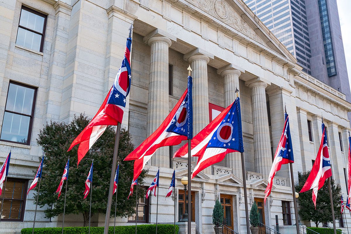 Multiple Ohio state flags are displayed in front of a large stone government building with tall columns and a modern skyscraper visible in the background.