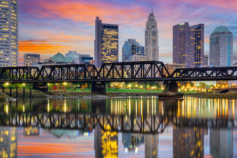 Downtown city skyline with modern buildings, a railroad bridge, and colorful sunset sky reflected in a calm river.