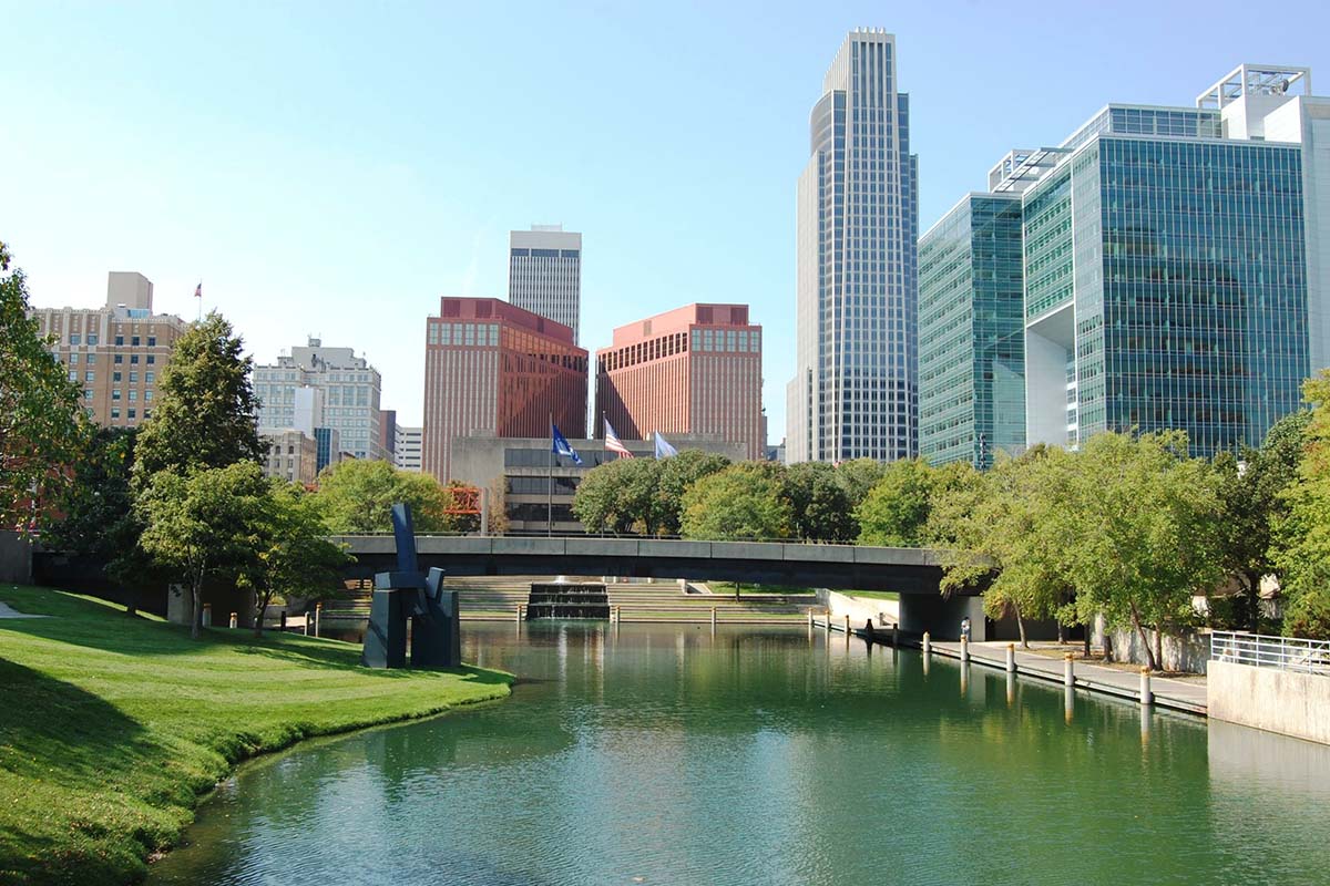 A river flows under a bridge with modern and historic office buildings in the background and trees lining both sides, on a sunny day.