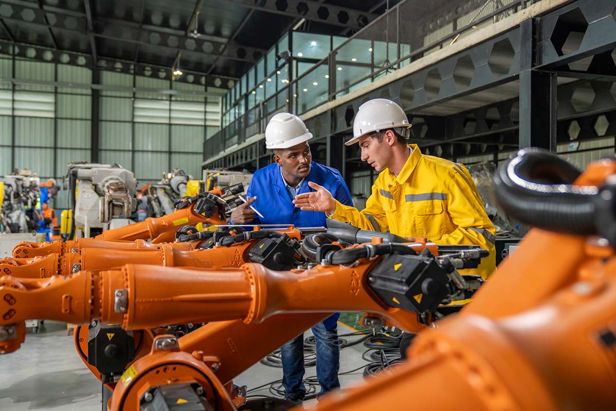Two workers in hard hats and safety gear discuss beside a row of orange industrial robotic arms inside a factory setting.