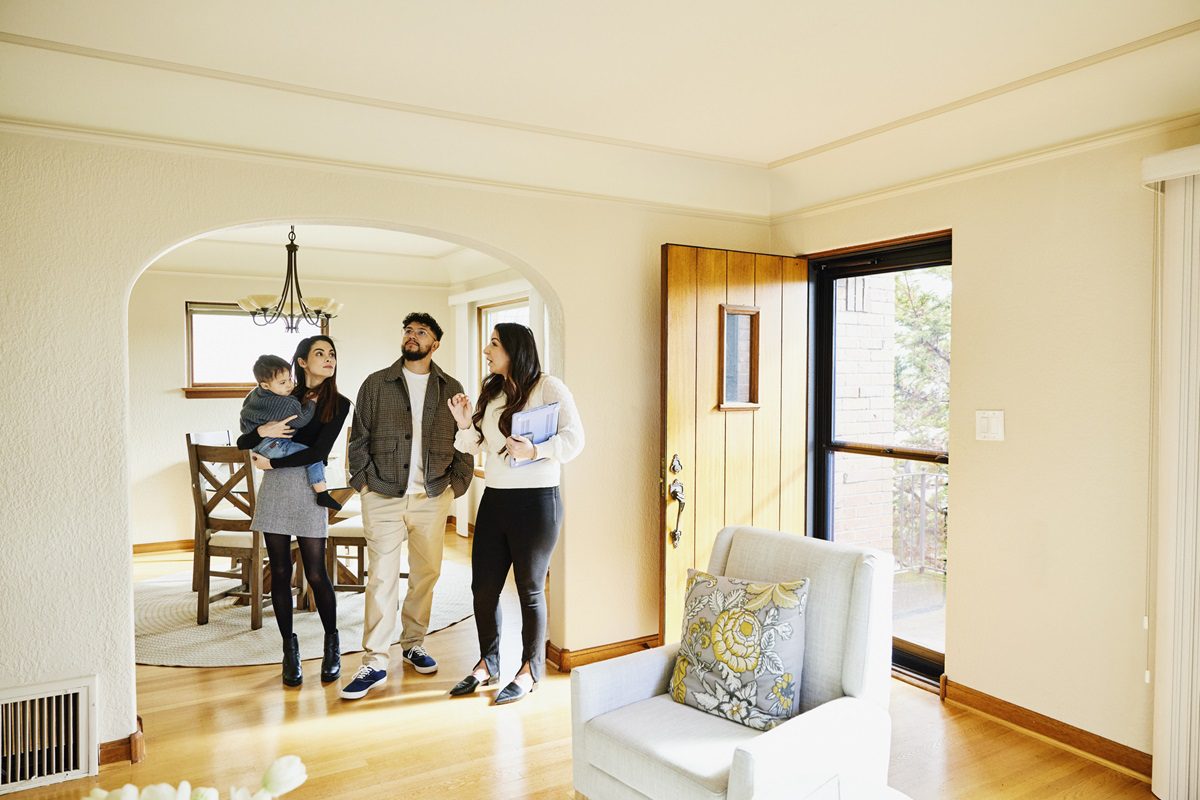 A couple holding a child tours a bright living room with a real estate agent, who is holding a clipboard.